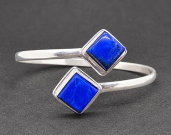 May include: A silver-toned open cuff bracelet featuring two square-shaped, deep blue lapis lazuli stones set at each end. The metal band has a smooth, polished finish, and the stones are framed in a simple, clean metal setting.