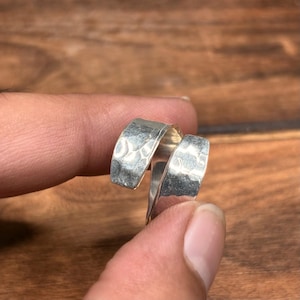 Puede incluir: Un anillo de plata tipo bypass con un acabado martillado y texturizado en la superficie exterior. La banda está abierta, con dos extremos que se superponen creando un diseño envolvente. El metal tiene un aspecto pulido con hendiduras.