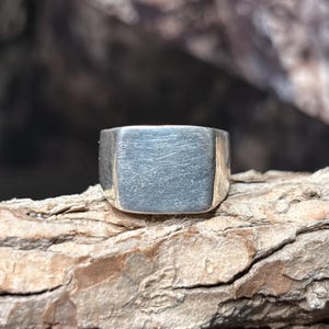 May include: A silver signet ring with a rectangular, flat face and a brushed, matte finish. The metal surface shows fine, horizontal scratch-like textures across the front, creating a muted, non-reflective appearance on the band.