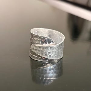 May include: A wide silver-toned ring featuring an overlapping wrap-around design. The surface of the metal is textured with a repeating pattern of small, rounded scales or indentations that cover the entire exterior of the band.