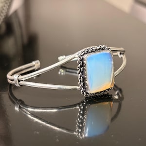 May include: A silver-toned cuff bracelet featuring a rectangular, milky white opal-like stone set in a decorative, textured metal frame. The band consists of two thin, smooth silver-toned wires that meet at the sides to support the central stone setting.