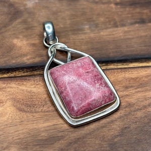 Puede incluir: Un colgante rectangular de rodonita con una piedra rosa moteada y pulida engastada en un marco de plata liso. El colgante incluye una reasa de plata en la parte superior, y la piedra muestra vetas blancas naturales y una textura superficial.