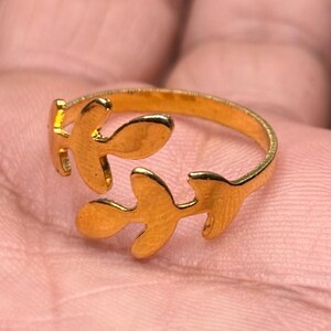May include: A gold-toned open ring featuring two symmetrical leaf-shaped motifs that meet in the center. The band is thin and circular, with the two leaves extending from opposite sides of the ring to create an adjustable, bypass-style design.
