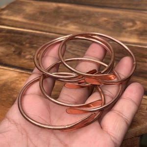 Könnte beinhalten: Ein Set aus drei dünnen, kreisförmigen Kupferarmreifen. Jeder Armreif hat eine glatte, metallische Oberfläche mit einem warmen rotbraunen Farbton. Die Armreifen sind offen gestaltet und laufen an den Enden in abgerundete Spitzen aus.