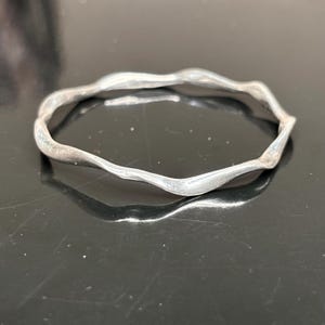 May include: A silver-toned bangle bracelet featuring a continuous, undulating, wavy design. The metal surface has a smooth, polished finish that reflects light along its irregular, curved edges, creating a fluid and organic circular shape.