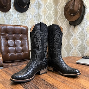May include: A pair of black leather cowboy boots featuring a textured crocodile-skin pattern on the foot and a tall shaft with intricate tonal stitching. The boots have a stacked wooden heel, a rounded toe, and pull tabs at the top of the shaft.