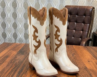May include: A pair of knee-high cowboy boots featuring a cream-coloured leather base with brown suede overlays. The boots have a pointed toe, a stacked wooden heel, and decorative brown suede cutouts along the shaft and collar.