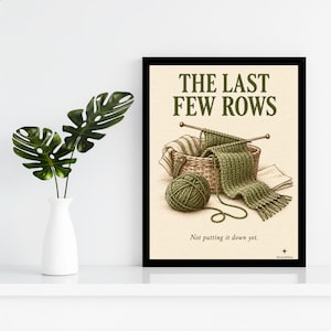 May include: A framed wall art print featuring a basket of green yarn, knitting needles, and a partially knitted scarf. The text reads THE LAST FEW ROWS and Not putting it down yet. The illustration is rendered in shades of green and beige.