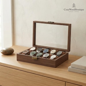 Puede incluir: Una caja expositora de madera oscura de forma rectangular con tapa de cristal transparente y cierre metálico. El interior cuenta con diez compartimentos individuales, cada uno con un reloj de pulsera con diferentes esferas.