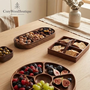 May include: A collection of dark brown wooden serving trays including an oval three-section tray filled with nuts and dried fruit, a square four-section tray with a handle containing crackers, and a round multi-section tray holding fresh fruit.