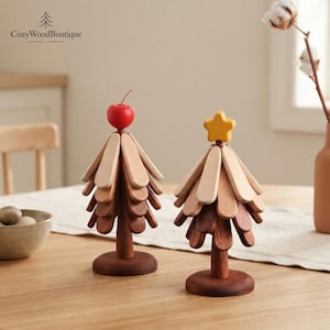 May include: Two decorative wooden tree sculptures with tiered, rounded branches in varying shades of brown. One tree is topped with a small red apple, and the other features a yellow star. Both stand on circular bases, crafted from smooth wood.