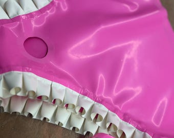 May include: A glossy pink garment featuring white ruffled trim along the edges and a circular cutout detail. The material has a smooth, reflective surface, and the design includes layered white ruffles and a thin pink ribbon accent.
