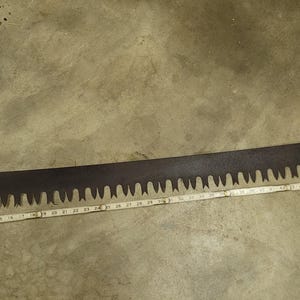 May include: A long, dark metal crosscut saw with two wooden handles attached at each end. A white measuring tape is positioned along the length of the blade, indicating a total span of approximately 60 inches from handle to handle.
