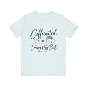 May include: A light heather blue short-sleeve t-shirt featuring the dark green text Caffeinated and Doing My Best with a graphic of a coffee cup and small pink hearts printed on the center of the chest in a script and sans-serif font.