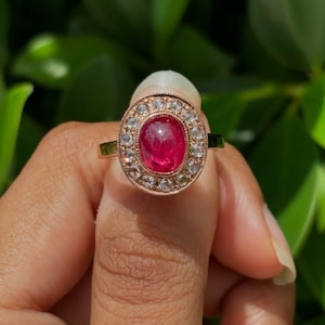 Könnte beinhalten: Ein Ring aus Roségold mit einem zentralen, ovalen, im Cabochon-Schliff geschliffenen roten Edelstein, der von einem Kranz aus kleinen, runden, klaren facettierten Steinen umgeben ist. Die Fassung hat einen Milgrain-Rand und die Ringschiene ist poliert.
