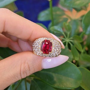 May include: A gold ring featuring a large, rectangular, deep red gemstone at the center, surrounded by a dense pavé setting of numerous small, clear, sparkling white stones that cover the entire wide band of the jewelry piece.