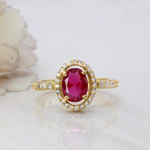 Pode incluir: Um anel de ouro com uma pedra preciosa central de corte oval em rosa escuro, rodeada por um halo de pequenas pedras claras. A banda também está cravejada com pequenas pedras claras nas laterais, criando uma aparência brilhante e reflexiva.