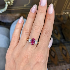Könnte beinhalten: Ein Ring mit drei Steinen, bestehend aus einem zentralen, oval geschliffenen, leuchtend pink-roten Edelstein, der von zwei kleineren, ovalen, klaren weißen Steinen flankiert wird. Die Steine sitzen auf einer dünnen, polierten Ringschiene aus Gelbgold.