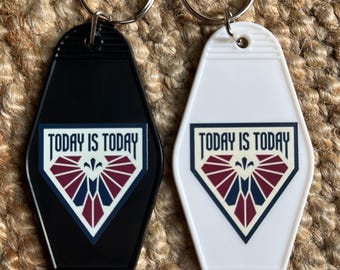 Peut inclure: Deux porte-clés de style motel, un noir et un blanc, chacun doté d'un anneau en métal. Le centre de chaque porte-clés présente un motif en forme de bouclier avec le texte "TODAY IS TODAY" au-dessus d'un design géométrique bordeaux et bleu.