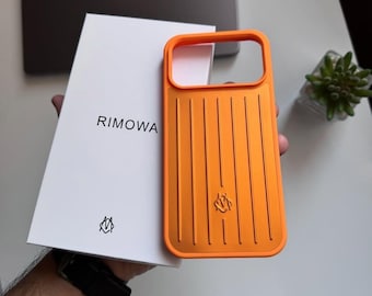 Puede incluir: Una funda de teléfono de color naranja brillante con una textura de ranuras verticales y un pequeño logotipo, presentada junto a su caja de embalaje rectangular blanca. La caja muestra el nombre RIMOWA en negro con un pequeño logotipo.