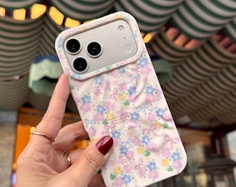 Puede incluir: Una funda para smartphone de color rosa claro con un patrón repetitivo de pequeñas flores azules, moradas y amarillas intercaladas con delicados lazos rosa claro. La funda tiene un marco elevado para la cámara y una superficie texturizada.