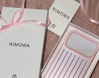 Puede incluir: Una funda para teléfono de color rosa claro y plateado con un patrón de ranuras verticales, presentada en una caja blanca con el logotipo de Rimowa y una etiqueta a juego, atada con una cinta rosa claro. La funda tiene un recorte rectangular.