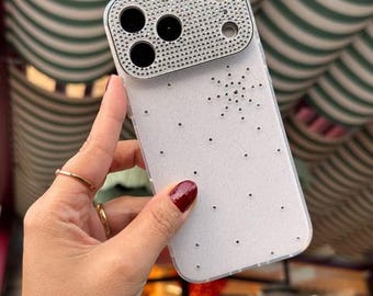 Puede incluir: Una funda transparente para smartphone con un módulo de cámara rectangular cubierto de pequeños brillantes plateados reflectantes. La parte posterior de la funda está decorada con un patrón disperso de diminutos brillantes y un diseño de estrella.