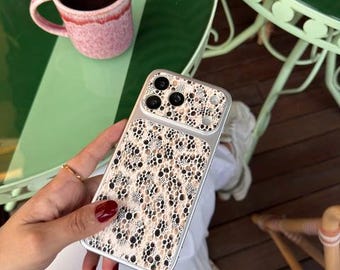 Puede incluir: Una funda para smartphone con un estampado animal en beige y negro con una superposición texturizada similar al encaje. La funda tiene un borde transparente y una sección recortada rectangular para las lentes de la cámara.