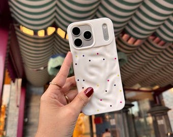 Puede incluir: Una funda de smartphone blanca con textura ondulada y pequeños puntos multicolores dispersos. La funda presenta un área de recorte rectangular elevada para la cámara y un acabado mate suave, diseñada para un teléfono con sistema de triple lente.