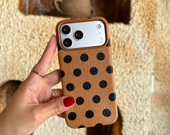 Puede incluir: Una funda de teléfono color canela con un patrón repetitivo de lunares negros. La funda presenta un recorte rectangular para la cámara con un borde canela, revelando un módulo plateado con tres lentes circulares y un flash.
