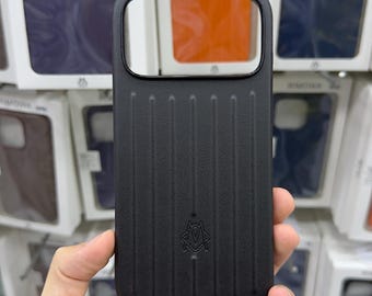 Puede incluir: Una funda de smartphone negra con textura de ranuras verticales y un pequeño logotipo en relieve en la parte inferior central. La funda presenta un gran recorte rectangular en la parte superior para la lente de la cámara sobre un fondo de cajas.