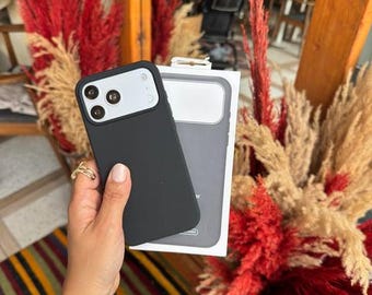 Puede incluir: Una funda de smartphone en color negro mate con un área de recorte blanca para la cámara, junto a su embalaje de venta blanco. La funda presenta una abertura para triple lente y una superficie trasera lisa y de color sólido.
