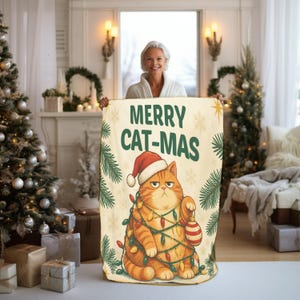 May include: A cream-colored throw blanket featuring a graphic of an orange tabby cat wearing a red Santa hat, tangled in colorful Christmas lights and holding a striped ornament. The text MERRY CAT-MAS is printed in green above the cat.