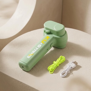 May include: A sage green handheld string launcher device with the text KU-STRING printed in yellow on the handle. The set includes a bright neon yellow string and a white USB charging cable, all presented as a complete electronic gadget set.