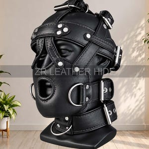 Peut inclure: Un masque de tête en cuir noir doté de multiples sangles réglables, de boucles, de rivets et d'anneaux en D en métal argenté. Le masque présente une ouverture pour la bouche et est posé sur un mannequin. Le texte ZR LEATHER HIDE est visible.