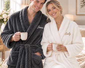 Op de afbeelding: Een donkergrijze pluche badjas met de naam James in het wit geborduurd en een witte pluche badjas met de naam Emily in het bruin geborduurd. Beide badjassen hebben een sjaalkraag, lange mouwen, een ceintuur en twee voorzakken.