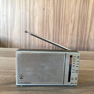 Könnte beinhalten: Ein tragbares Grundig Boy 30 Radio mit silbernem Metall-Lautsprechergitter, schwarzem Gehäuse und ausziehbarer Antenne. Die Frontseite zeigt eine vertikale Frequenzskala für AM- und FM-Bänder sowie das Grundig-Logo und ein kleines Wappen.