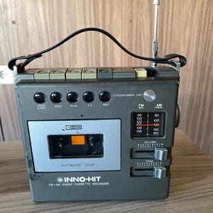 Puede incluir: Una grabadora de radio casete FM-AM Inno-Hit de color gris con asa de transporte superior, antena telescópica y compartimento para casetes frontal. El dispositivo incluye botones de control, un dial de sintonización y deslizadores de volumen.