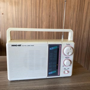 Może przedstawiać: Białe przenośne radio INNO-HIT AM/FM 2-pasmowe z górnym uchwytem i wysuwaną metalową anteną. Z przodu znajduje się kratka głośnika, panel wyświetlacza częstotliwości AM i FM, dwa okrągłe pokrętła do strojenia i głośności oraz przełącznik.