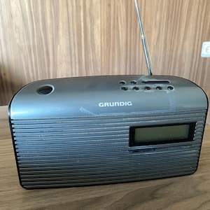 Könnte beinhalten: Ein graues Grundig-Radio mit einer horizontal gerippten Frontblende, einem digitalen Display, einer ausziehbaren Metallantenne an der Oberseite und mehreren Bedientasten. Das Gerät hat eine schlanke, abgerundete rechteckige Form.
