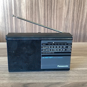 May include: A black Panasonic portable radio with a silver telescoping antenna. The front panel features a speaker grille, a frequency tuning scale for FM-UKW and AM-MW bands, and text reading AC-BATTERY BUILT IN AC TRANSFORMER.