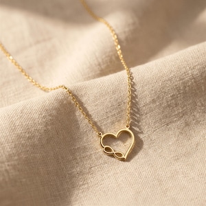 May include: A gold-toned necklace featuring a delicate chain and a pendant shaped like an open heart outline. The bottom left corner of the heart incorporates a small infinity symbol integrated into the frame of the heart shape itself.