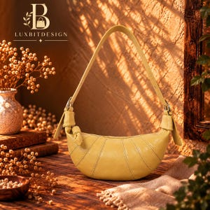 May include: A crescent-shaped yellow shoulder bag featuring a smooth finish, vertical stitched panel detailing, and a single adjustable strap attached with silver-toned metal hardware. The bag has a structured, curved silhouette with knotted accents.