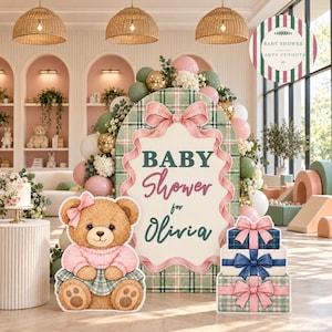 May include: A baby shower party cutout set featuring a large oval sign with a green plaid pattern, a pink ribbon border, and the text Baby Shower for Olivia. Includes a teddy bear cutout in a pink sweater and three stacked gift boxes.