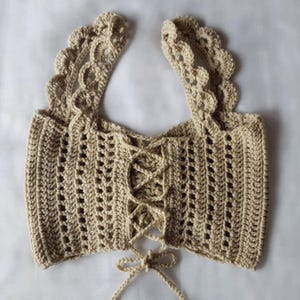 May include: A beige crochet crop top featuring a lace-up front closure with matching string ties at the bottom. The garment has wide, scalloped shoulder straps and a textured, open-knit pattern throughout the bodice, creating a structured look.