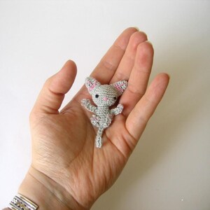 May include: A small, light gray crocheted cat figurine featuring tiny pink stitched details on the ears and nose, with black bead eyes. The miniature animal has outstretched arms and legs, a long tail, and is crafted with a visible yarn texture.