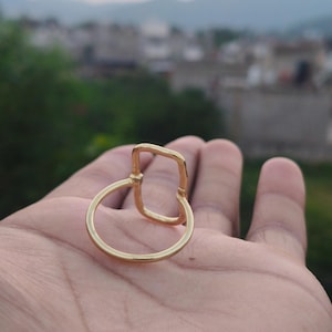 Könnte beinhalten: Ein goldfarbener Ring mit einem kreisförmigen Band, das mit einem vertikalen, rechteckigen Element verbunden ist. Das Metall hat eine glatte, polierte Oberfläche und das Design besteht aus einer einfachen, durchgehenden drahtartigen Struktur.