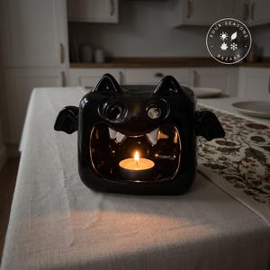 Puede incluir: Un portavelas de cerámica negra brillante con forma de murciélago, boca abierta, dos orejas puntiagudas y pequeñas alas laterales. Una vela de té encendida se encuentra en el centro hueco. El texto Four Seasons Bazaar es visible.