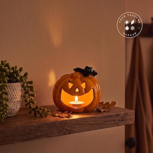 Peut inclure: Un bougeoir en céramique orange brillant en forme de citrouille d'Halloween avec un chat noir perché sur le dessus, des yeux triangulaires découpés et une large bouche contenant une bougie chauffe-plat allumée, avec des feuilles décoratives.