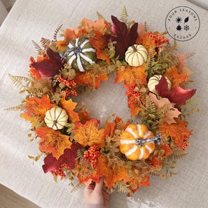 May include: A circular autumn wreath featuring orange, yellow, and deep red maple leaves, small white and orange pumpkins, and clusters of orange berries. The wreath includes green fern-like foliage and a logo reading Four Seasons Bazaar.